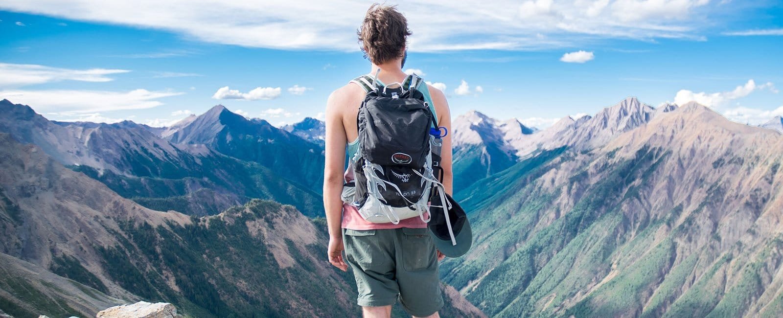 Persona que se encuentra mirando las montañas. persona que esta en la cima de la montaña apreciando la vista trae kuna mochila de color negro con gris y una gorra colgando en la mochila, trae un termo de agua.