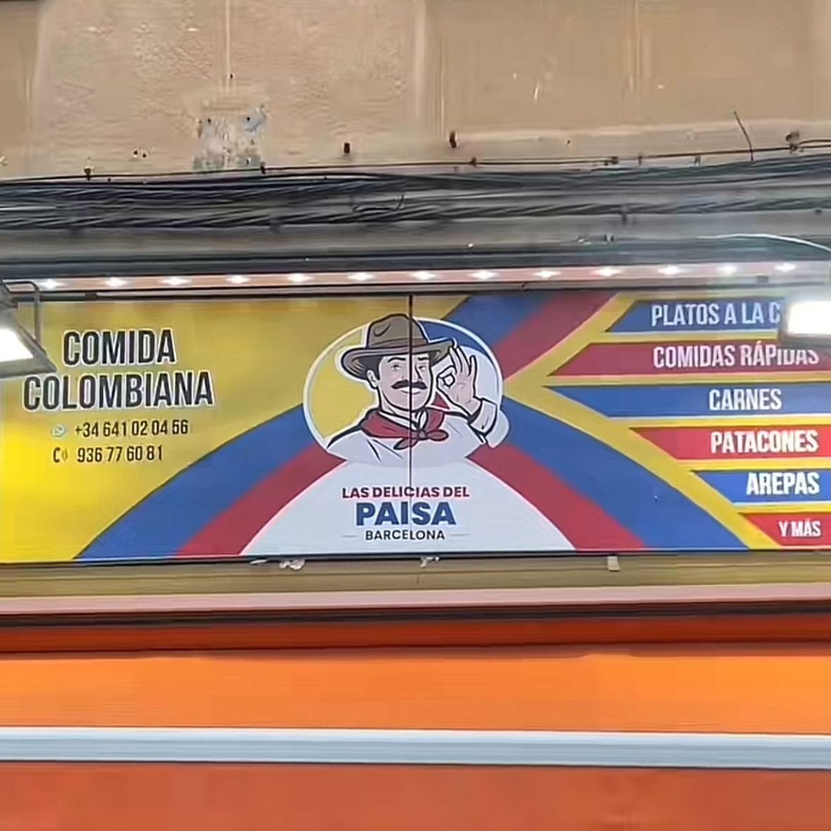 Cartel colorido de una cocina de comida colombiana, ubicado en Barcelona. En el cartel se puede ver parte de la lista de platillos que contiene su carta y su número de contacto. Se ve una persona animada, con semblante feliz y satisfecho, con vestimenta tradicional de las ciudades o pueblos de Latinoamérica. en la parte de arriba del cartel y también a los lados se pueden ver lámparas que iluminan el mismo cartel y parte de la entrada del lugar, el cual aparentemente tiene un color muy vistoso y atractivo como lo es el naranja. Por encima del cartel hay un cableado, y más arriba se puede ver parte del edificio, en colores cálidos como el beige o marrón.