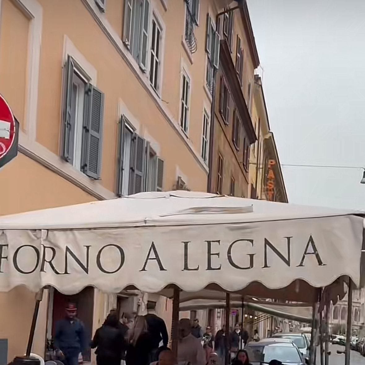 Cocina o restaurante de comida italiana, a unos metros del Coliseo de Roma, con un ambiente al aire libre. El cielo nublado da una señal del clima frío y la zona parece ser residencial, pues se ven edificios con ventanas rectangulares. Hay una carpa de color crema con la frase “Forno a Legna” a los costados, debajo de ella hay mesas y sillas. Hay personas comiendo debajo de la carpa, pero también hay gente caminando sobre la banqueta, todos con abrigo y chaquetas, cubriéndose del frío a causa de la baja temperatura.