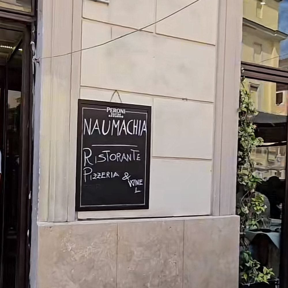 Se ven la entrada delantera de un restaurante o cocina y en la parte delantera del establecimiento se ve un cartel con el nombre del restaurante. El edificio tiene una fachada simple, parece ser un diseño arquitectónico minimalista.