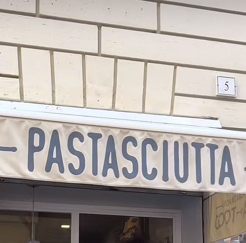 Se miran unas letras en mayúsculas con el texto "Pastasciutta", que es una cocina de pastas, está en la parte baja de un edificio y en la entrada se ve una puerta de cristal.