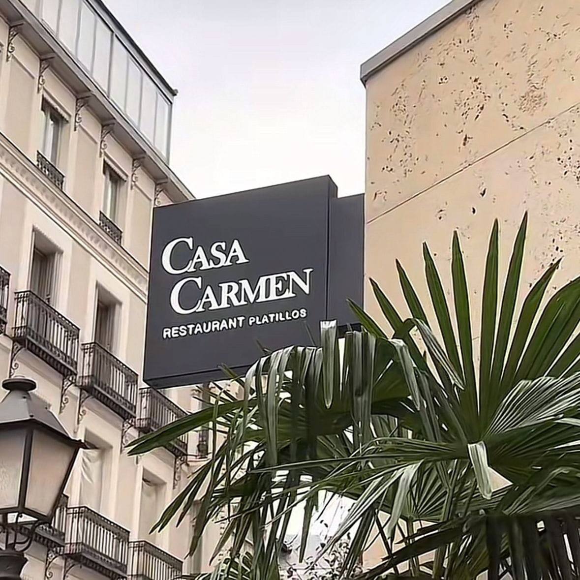 Restaurante Casa Carmen, en la esquina de una calle de Madrid, concretamente en una zona residencia. En la imagen se ve un letrero del restaurante de comida, con unas letras blancas que contrastan con el fondo negro. Se ve una palmera al lado del establecimiento y una lámpara cerca de la palmera, La arquitectura tradicional del edificio de enfrente indica una construcción clásica, También se ven unos balcones con barandal. El clima parece ser frío por el cielo nublado. Los mejores restaurantes de Europa