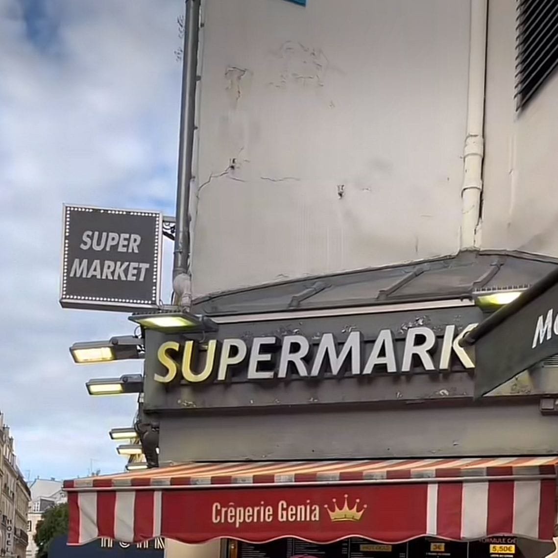 Se ve un local de comida rápida o postres justo en la parte baja de un edificio, al lado de una tienda de productos o un Supermarket. Las letras del "Supermarket" son grandes y llamativas por el color que tienen, sin embargo, la crecería es pequeña y solo dan la comida para llevar, y se puede ver que es una zona residencial en donde está ubicada. El cielo parece estar nublado, lo que podría causar un clima frío.