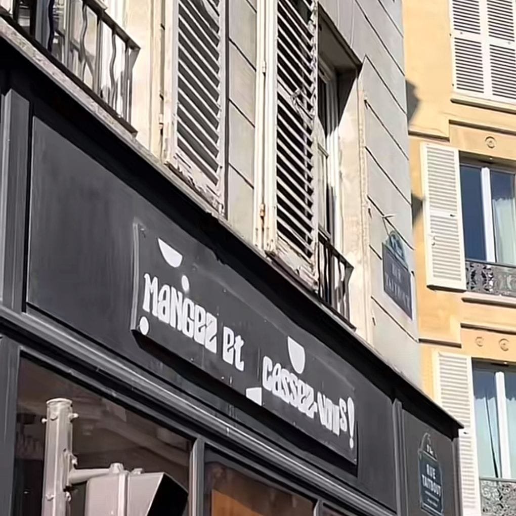 Fachada de un edificio urbano de arquitectura europea, en ella hay un letrero negro con letras blancas y tipografía moderna que dice "Mangez Et Cassez Vous" y hay elementos gráficos como puntos y semicírculos blancos que decoran el fondo negro. Los edificios tienen persianas tradicionales de madera pintadas de gris, con un estilo clásico y tienen ventanas con barandillas. La luz del sol indica un clima caluroso y en conjunto, la escena tiene un aire moderno y urbano mezclado con un entorno clásico europeo. Los mejores restaurantes de Europa