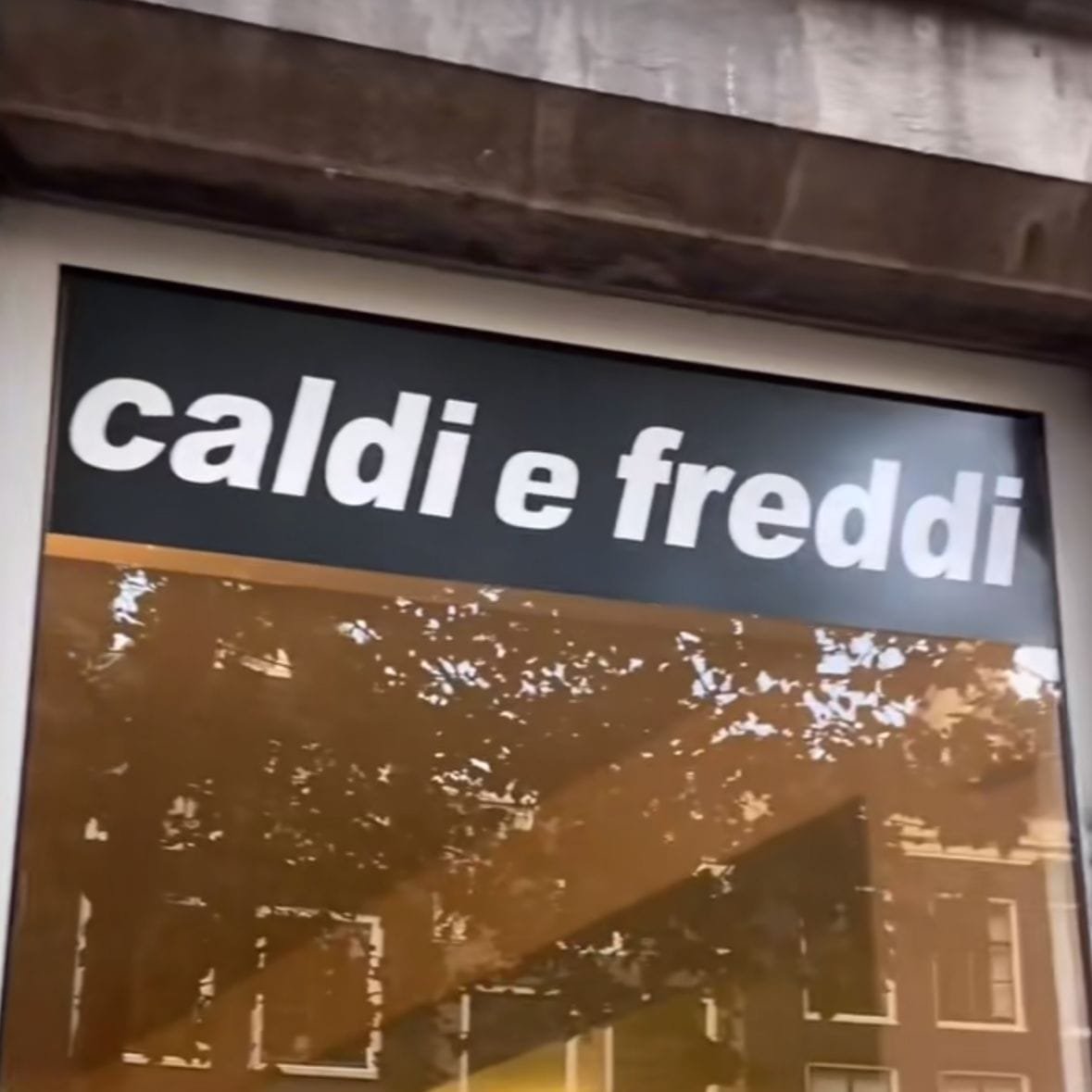Fotografía de la fachada de un restaurante o cocina local. Destaca un gran cartel arriba de una ventana o puerta de vidrio. El cartel tiene un fondo negro con texto en letras blancas que dice: "caldi e freddi" en italiano, se traduce al español como "calientes y fríos". El texto tiene una tipografía simple, en negrita y en minúsculas, lo que le da un aspecto moderno y claro, con un tono informal. En el vidrio se refleja el exterior y se distinguen los reflejos de árboles y edificios de ladrillo con ventanas rectangulares. Se perciben colores cálidos y tenues dentro del local y por la iluminación exterior se puede ver un clima templado o frío. En la parte superior de la imagen muestra el marco de la estructura del edificio, hecho de concreto.