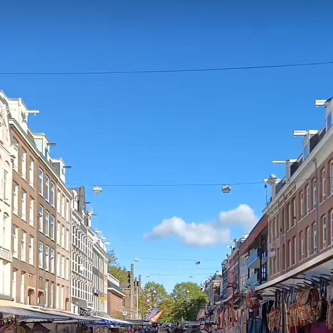Se aprecia un mercado al aire libre sobre la calle, en él venden comida, productos y muchas cosas más. También hay cafeterías, bares y restaurantes alrededor, ideales para sentarse y disfrutar el ambiente. Hay una mezcla de culturas, oír distintos idiomas, encontrar productos internacionales, y conocer vendedores de distintos orígenes. Parece ser una zona residencial, pues hay edificios de ladrillo, con balcones y fachadas altas. El cielo aquí parece estar despejado de nubes y la luz del sol por la mañana parece crear un clima cálido.