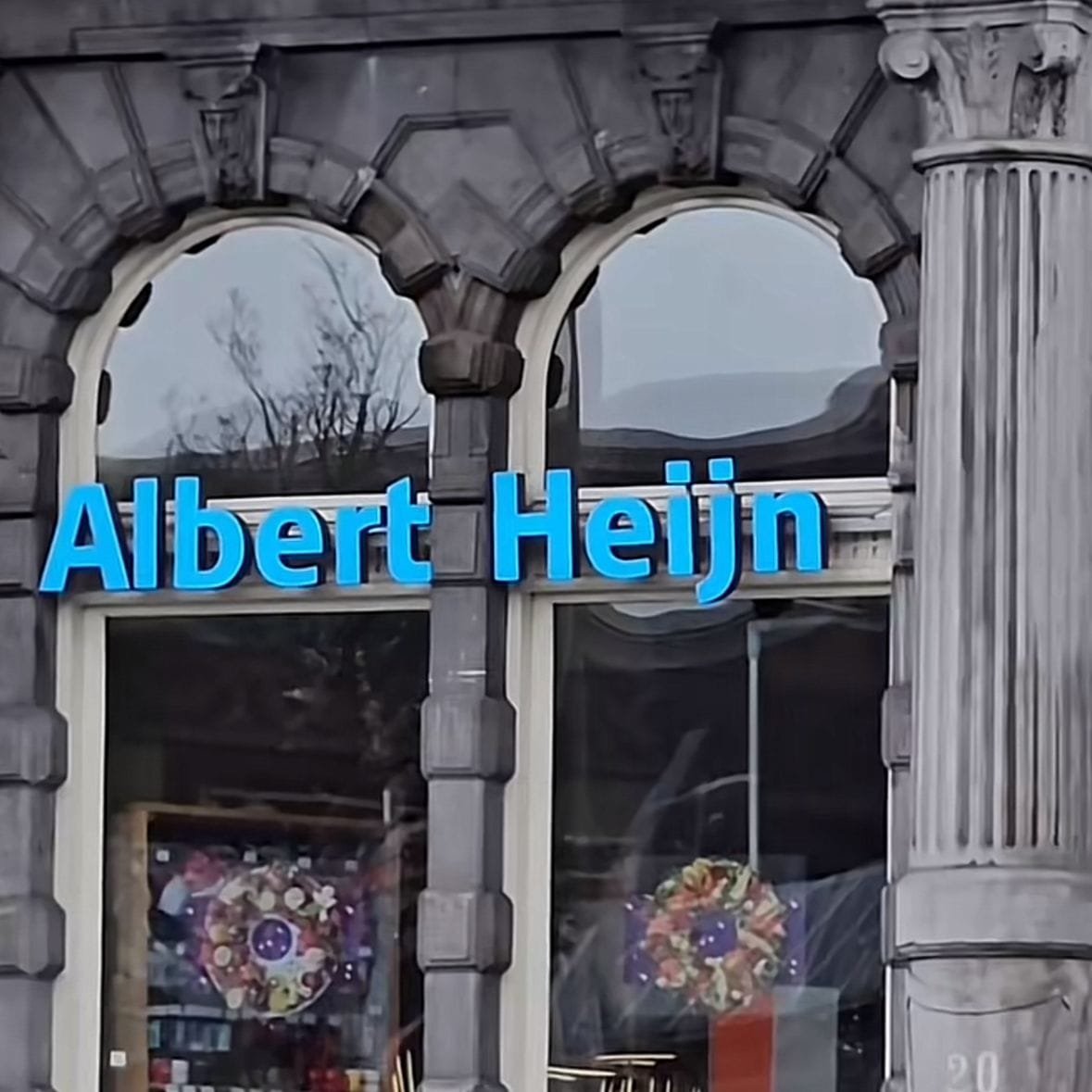 Se ve el nombre del establecimiento en letras color azul, sobre unos ventanales que permiten la vista directa al interior de la tienda, el edificio parece estar construido de concreto y por la textura parece ser algo antiguo, con un diseño arquitectónico un poco antiguo.Por la iluminación y el reflejo en los cristales, se puede asumir un clima frío.
