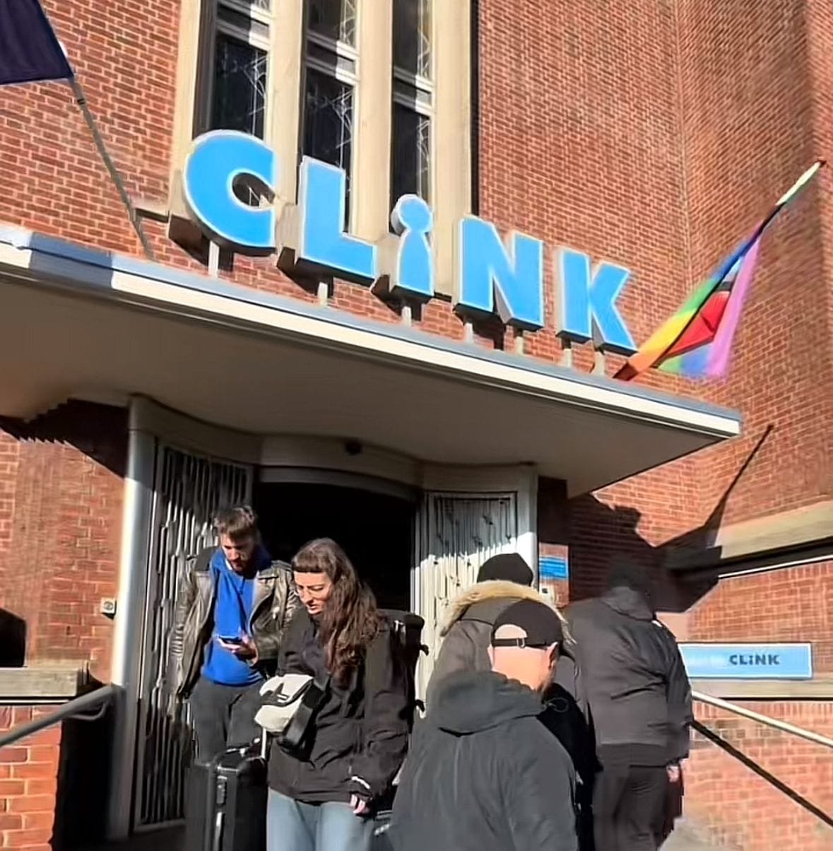 Hay personas saliendo y entrando del lugar, el edificio está hecho de ladrillo y tiene ventanas. Por la iluminación del sol se puede indicar que el clima es caluroso.