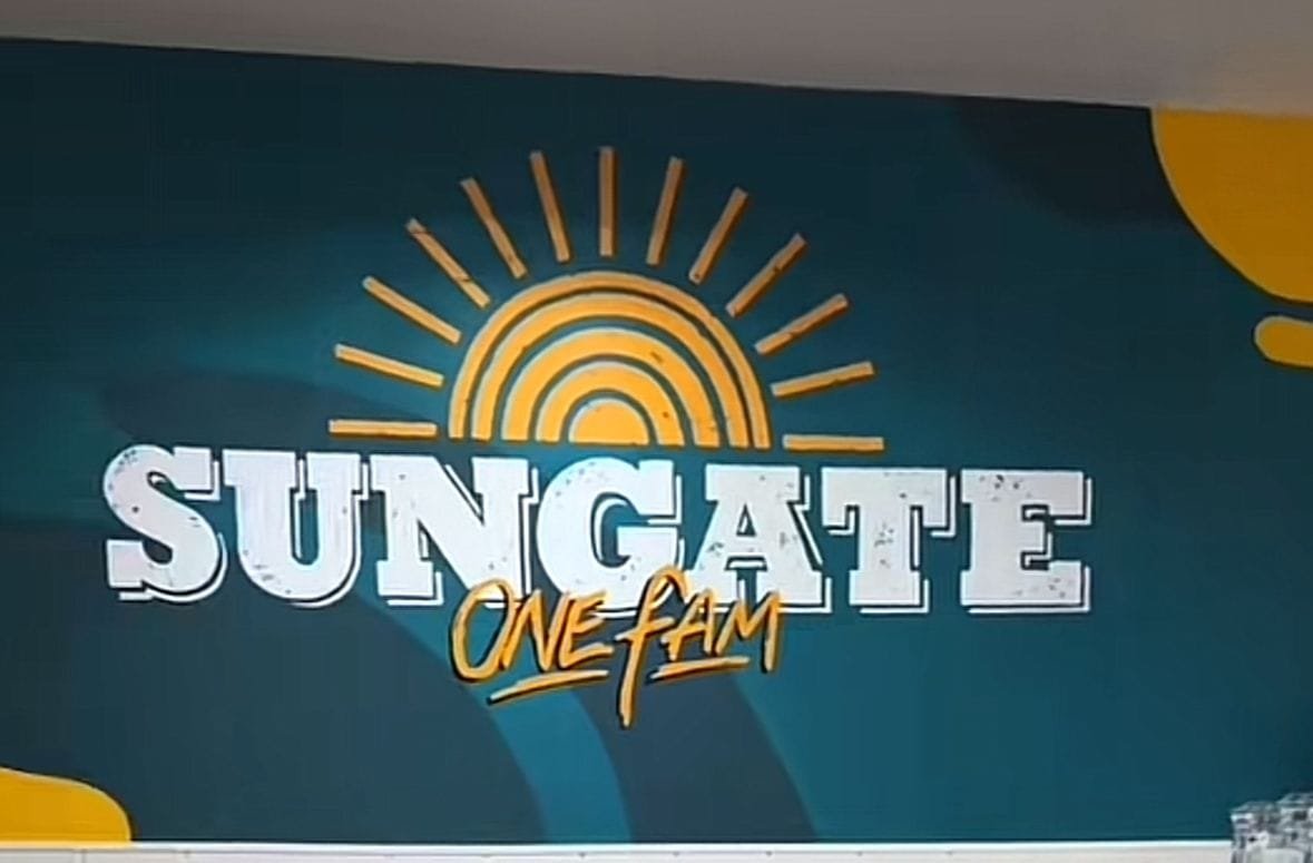 Mural de un hostal con un diseño llamativo y colorido. En el centro del mural se encuentra un texto grande que dice: "SUNGATE" y debajo, en un estilo informal y en color amarillo el texto: "ONE FAM". Encima de las letras se ve un sol en forma de semicírculo con líneas curvas en su interior que simulan rayos, y líneas rectas que irradian hacia arriba, todas en color amarillo. El fondo del mural es un color azul verdoso oscuro, el estilo da una sensación de energía, comunidad y calidez, la iluminación es cálida creando un ambiente acogedor y lleno de paz.