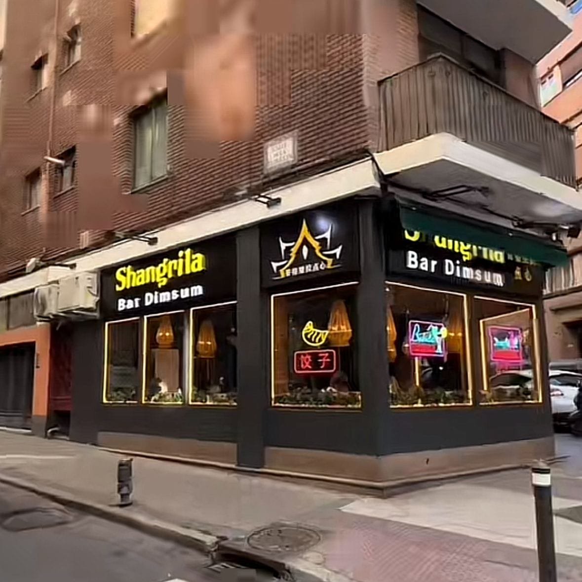 Aquí hay un restaurante con fachada negra y simple. Hay muchas ventanas con vista directa al interior del restaurante y sobre las ventanas se pueden ver letreros de luces de neón para llamar la atención de la gente que pasa por enfrente. Las letras con el nombre del restaurante son de colores llamativos y que contrastan con el fondo negro. El entorno es una zona residencial, se aprecian edificios de ladrillo con ventanas rectangulares, muy común en Madrid y Barcelona, y en el piso de arriba del restaurante se ve un balcón con barandal de metal. Los mejores restaurantes de Europa