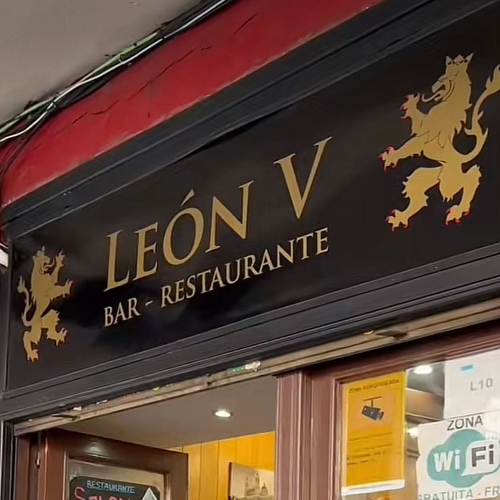 Bar/restaurante de nombre Leon V, se ve el nombre del establecimiento con letras doradas y unos leones en ambos lados del nombre. Las letras contrastan con su fondo de color negro y la fachada roja del edificio indica que quieren llamar la atención de sus clientes, lugar perfecto donde encontrar comida.