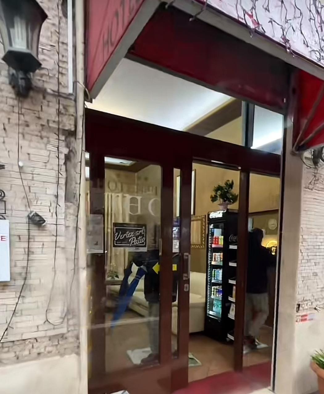 Entrada de un hotel, decorada con una domo rojo y puertas de vidrio. A través de las puertas, se puede ver un interior acogedor con un sofá blanco, una planta decorativa y una máquina de bebidas. Un letrero con estilo vintage está pegado al cristal.