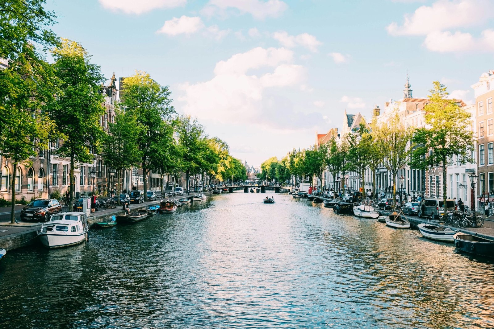 Canal de Ámsterdam durante el día. El agua refleja las fachadas estrechas y coloridas de las casas típicas holandesas. Bicicletas reposan junto a los puentes de hierro forjado, mientras pequeñas barcos y botes turísticos navegan por el canal. Árboles alineados a ambos lados ofrecen sombra y un toque natural al paisaje urbano. El ambiente es relajado, con gente paseando o simplemente disfrutando de la vista.
