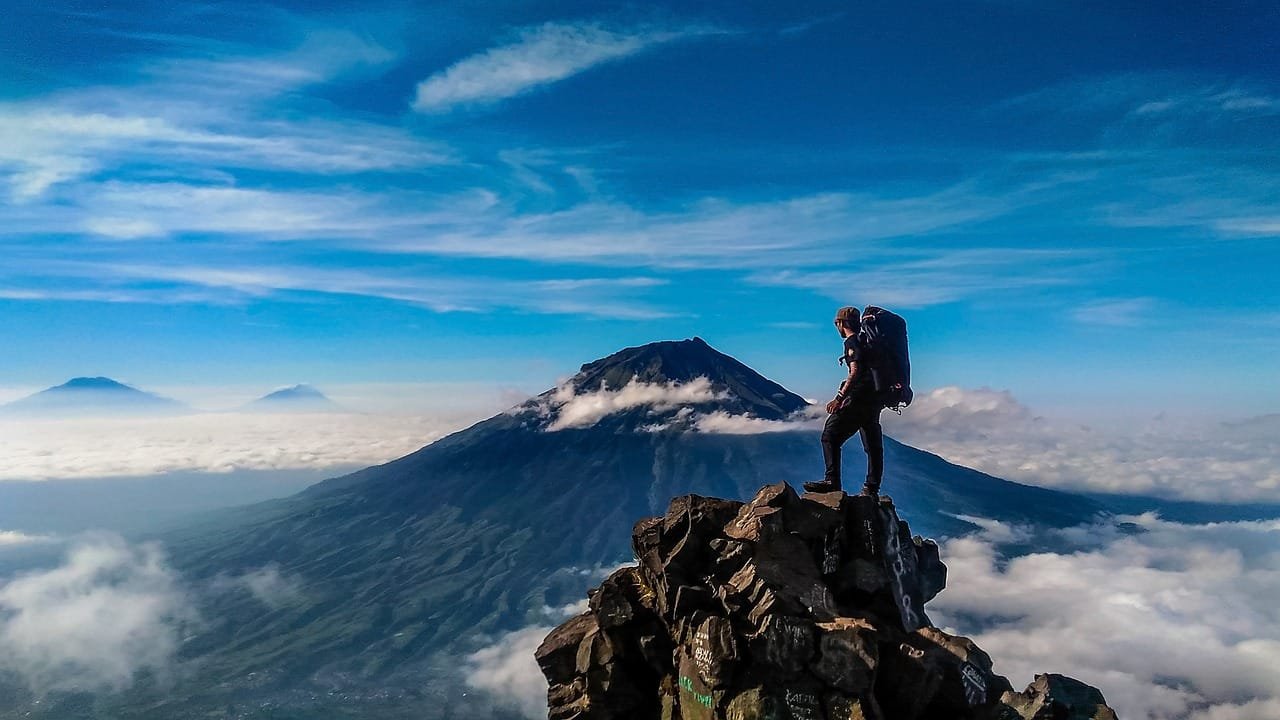 Una persona que es un excursionista solitario se encuentra en la cima de una montaña rocosa, admirando un paisaje impresionante de volcanes y nubes que cubren el valle