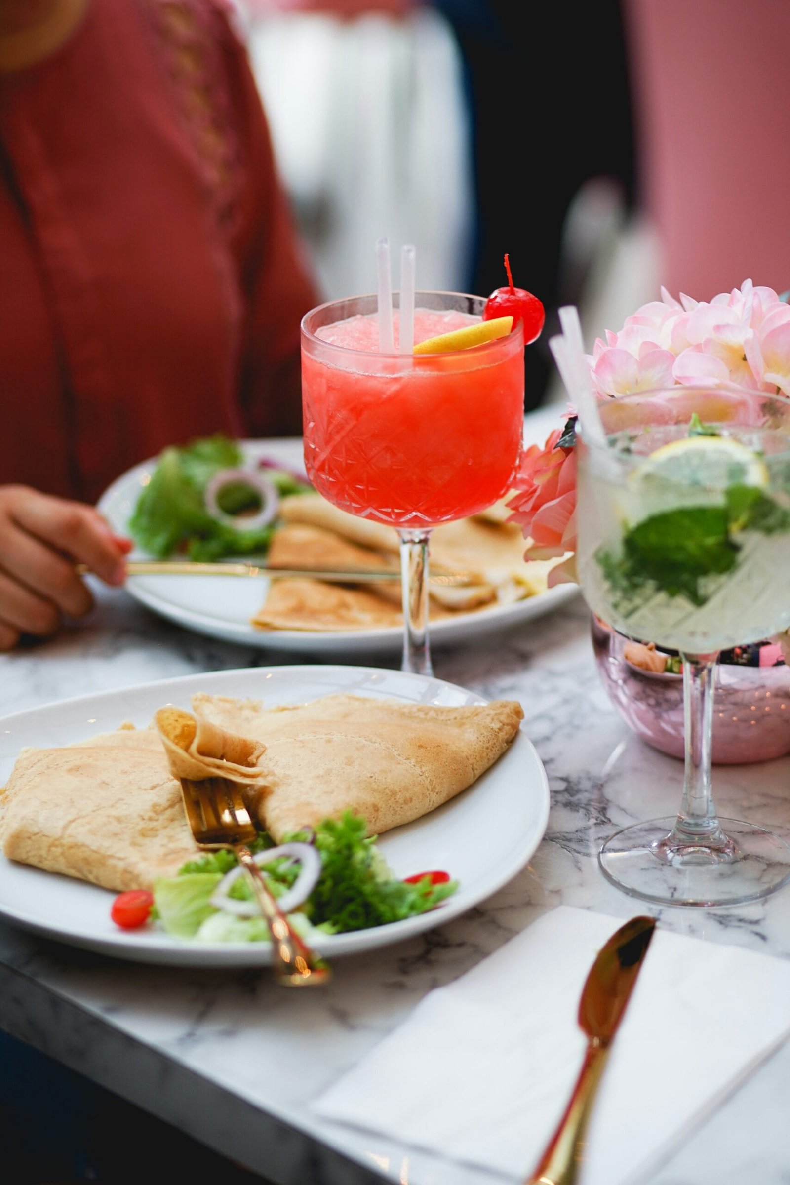 Comida en un restaurante o cafetería con ambiente elegante. Se ven platos con crepas, acompañadas de una ensalada de verdura. Se ven dos bebidas servidas en copa, los cubiertos son dorados y se ve una servilleta blanca, todo esto sobre una mesa de mármol. Se ve la silueta de una persona con camisa roja. Esta es una imagen donde se ve una comida ligera en un lugar bonito y elegante, de una salida entre amigos, la iluminación ayuda a crear un ambiente lleno de paz.