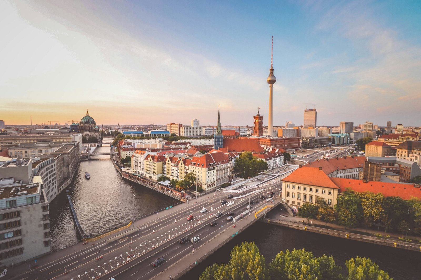 En la imagen se observa una vista panorámica de Berlín al atardecer, con el cielo teñido de tonos cálidos. Probablemente con un clima cálido. Destaca al centro la Torre de Televisión, símbolo moderno de la ciudad. Hay edificios históricos a los lados con tejados rojos y fachadas claras. Se observa una gran cúpula, que es de la Catedral de Berlín. Un río cruza la ciudad entre las construcciones, se miran puentes y barcos. El paisaje combina lo clásico con lo moderno, en un ambiente urbano, dinámico y lleno de historia.