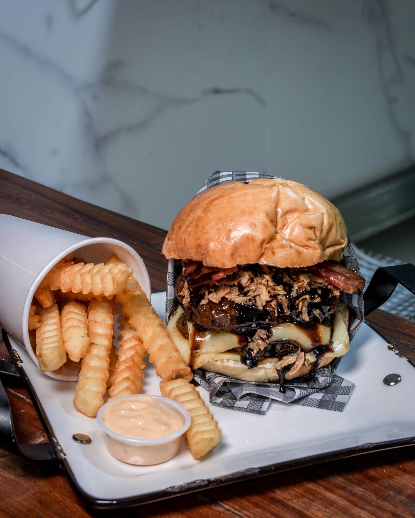 Deliciosa hamburguesa esponjosa de res con salchicha, con ingredientes como queso amarillo y salsa inglesa. En el lado izquierdo de la imagen se ve una porción acompañante de papas fritas con una forma muy tradicional y llamativa, también se ve un poco de aderezo, Todo esto sobre una charola, en un entorno muy higiénico y con la correcta iluminación para hacer que la comida se distinga muy bien. Parece que todo esto está sobre una barra de madera y de fondo se ve una pared blanca con líneas uniformes contrastantes.