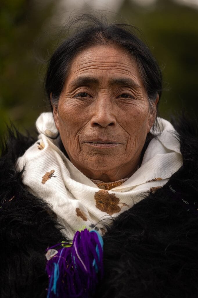 Una mujer de rostro sereno y mirada profunda, su expresión refleja sabiduría, fortaleza y una vida llena de experiencias, lleva una bufanda blanca con bordados de flores y una prenda negra de textura gruesa, adornada con hilos de colores.