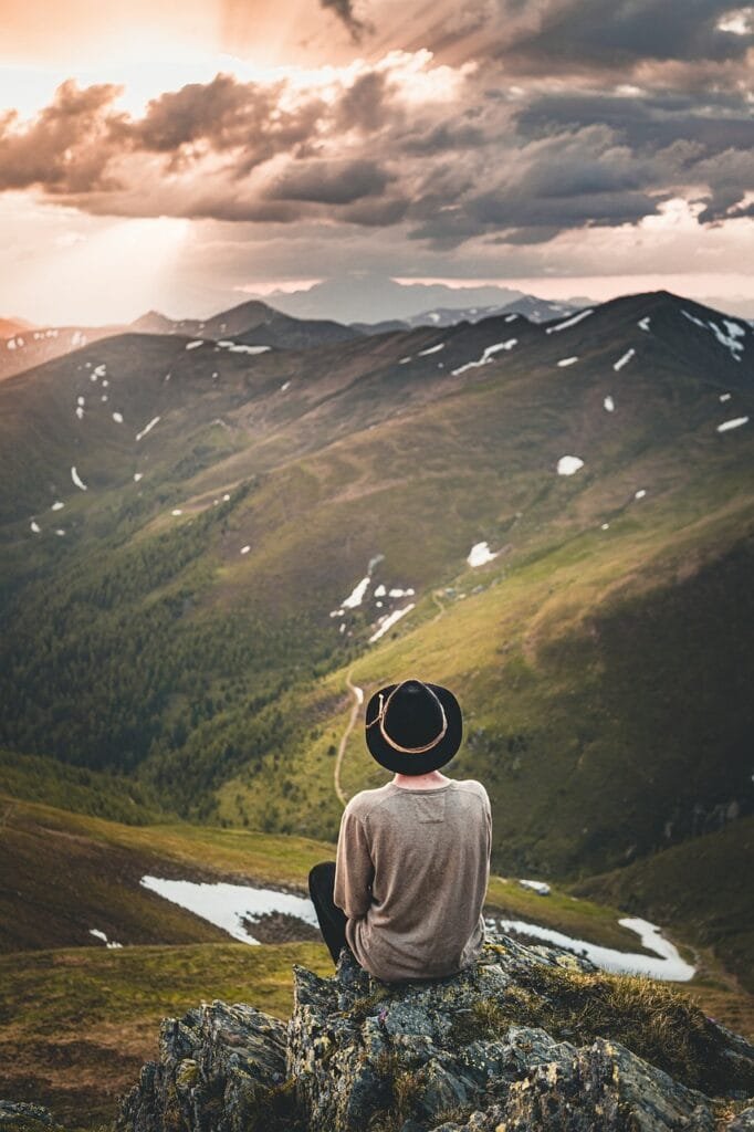 Persona sentada en la cima de una montaña contempla paisaje montañoso,lleva sombrero negro con una cuerda decorativa dorada y una playera beige, enfrente tiene un valle verde