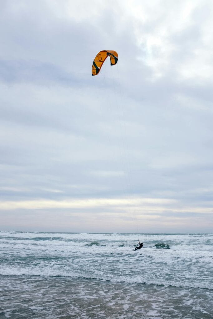 Persona practicando kitesurf en el mar durante un día nublado. El cometa de color naranja se eleva en el cielo mientras las olas golpean suavemente la orilla.
