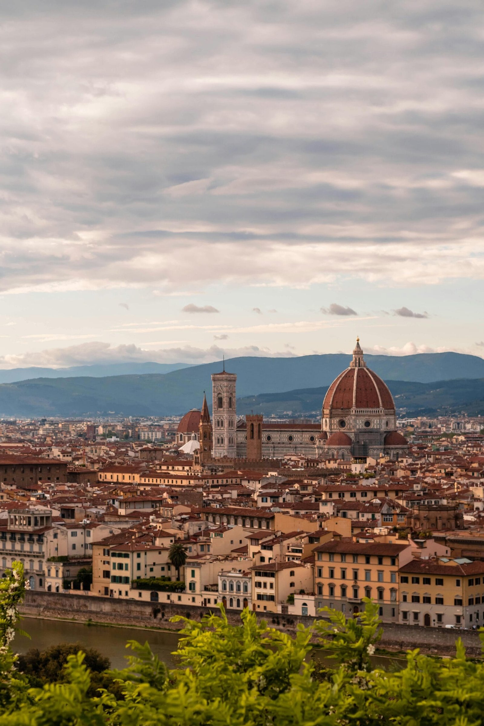 Paisaje urbano de Florencia Italia donde hay muchos tejados rojizos y destaca una Catedral con su gran cúpula de ladrillo. Al fondo, se observan unas colinas bajo un cielo parcialmente nublado. En primer plano, un río que refleja las fachadas históricas de los edificios que lo rodean. La escena combina arte, historia y belleza de un paisaje natural.