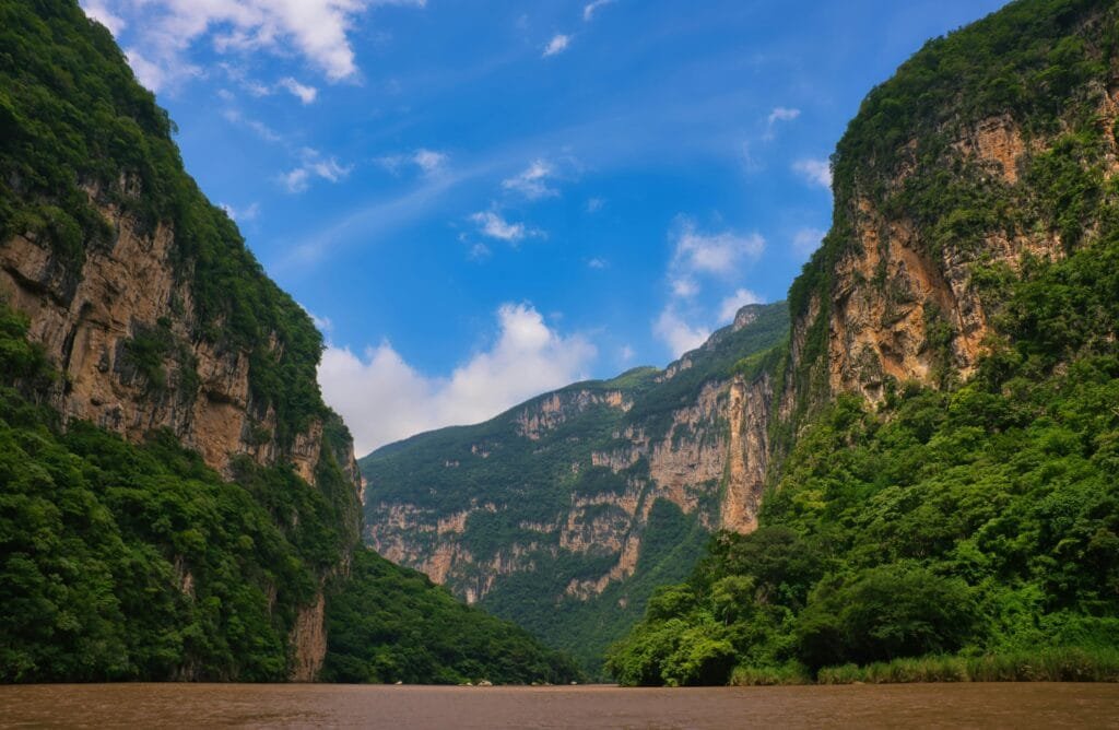 Un paisaje natural, un río tranquilo que se abre paso entre dos enormes paredes de roca, las montañas son altas, imponentes y están cubiertas parcialmente de vegetación, con un ambiente fresco, es un cañón profundo rodeado de naturaleza, un lugar que transmite grandeza