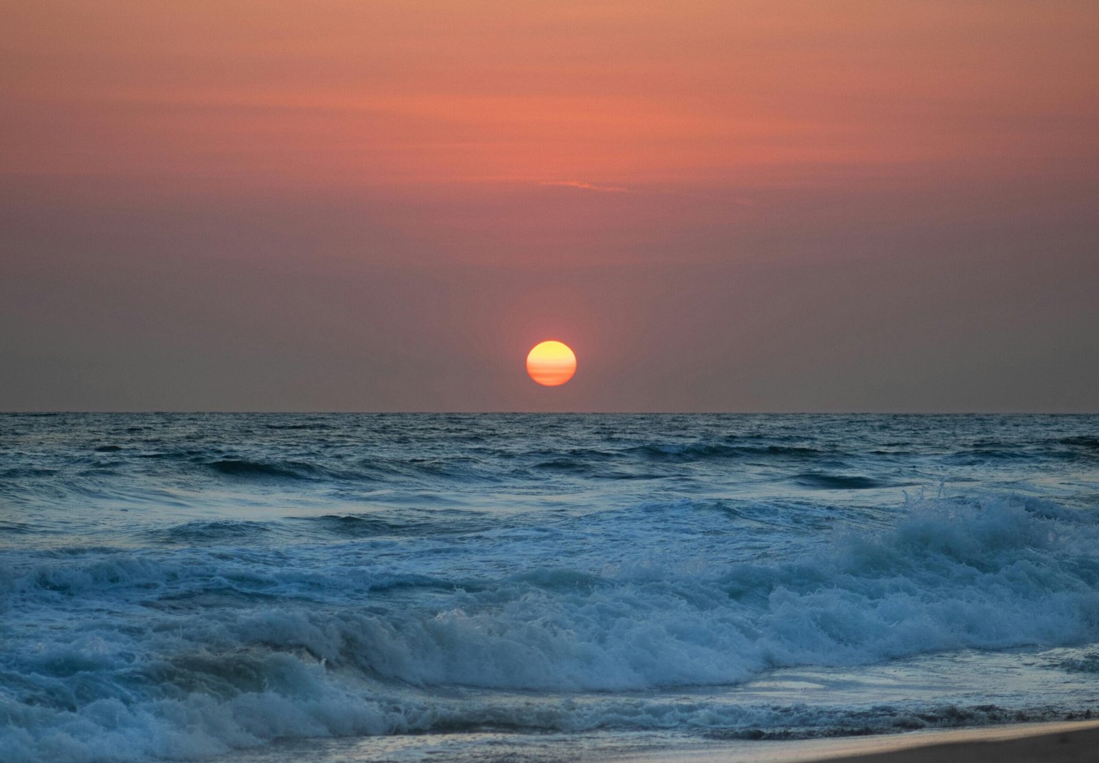 Una puesta de sol en la playa con el sol descendiendo en el horizonte, reflejando tonos cálidos de naranja y rosa sobre la arena húmeda y el mar en calma. Un paisaje sereno que evoca paz.