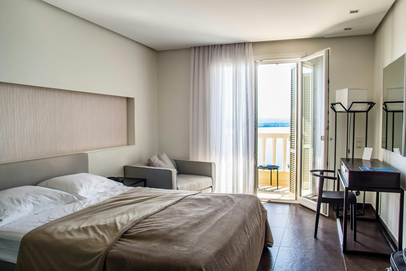 En la imagen se puede ver una habitación de hotel, lista para hacer uso de ella, pues está todo ordenado y muy limpio, la luz natural ayuda a crear un ambiente tranquilo y lleno de paz, así como el color de las paredes, cortinas, la cama y los muebles. En la habitación se ve lo necesario para dormir y relajarse, incluso para sentarse en el sofá, y al lado derecho se observa un escritorio, un espejo mediano y una lámpara. El clima o temperatura de la habitación parece ser neutral.