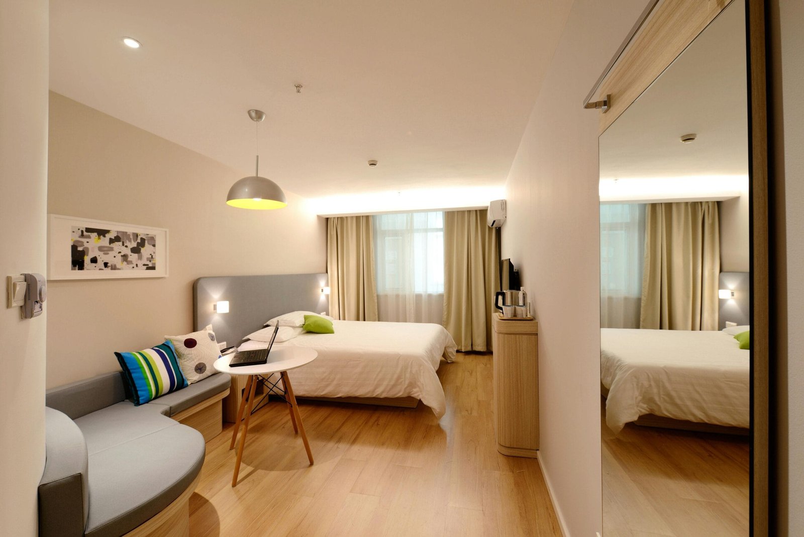 En la imagen se puede ver una habitación de hotel, lista para hacer uso de ella, pues está todo ordenado y muy limpio, las luces ayudan a crear un ambiente tranquilo y lleno de paz, así como el color de las paredes, cortinas, la cama y los muebles. En la habitación está lo indispensable para dormir y relajarse, incluso para trabajar sobre la mesa y el sofá que se encuentran al centro de la habitación, y al lado derecho se observa un espejo de gran tamaño, al fondo se aprecia un aire acondicionado para moderar el clima o temperatura de la habitación.