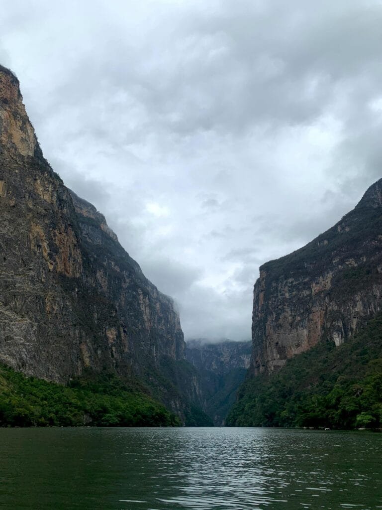 Un paisaje natural, un río tranquilo que se abre paso entre dos enormes paredes de roca, las montañas son altas, imponentes y están cubiertas parcialmente de vegetación, con un ambiente fresco, es un cañón profundo rodeado de naturaleza, un lugar que transmite grandeza.