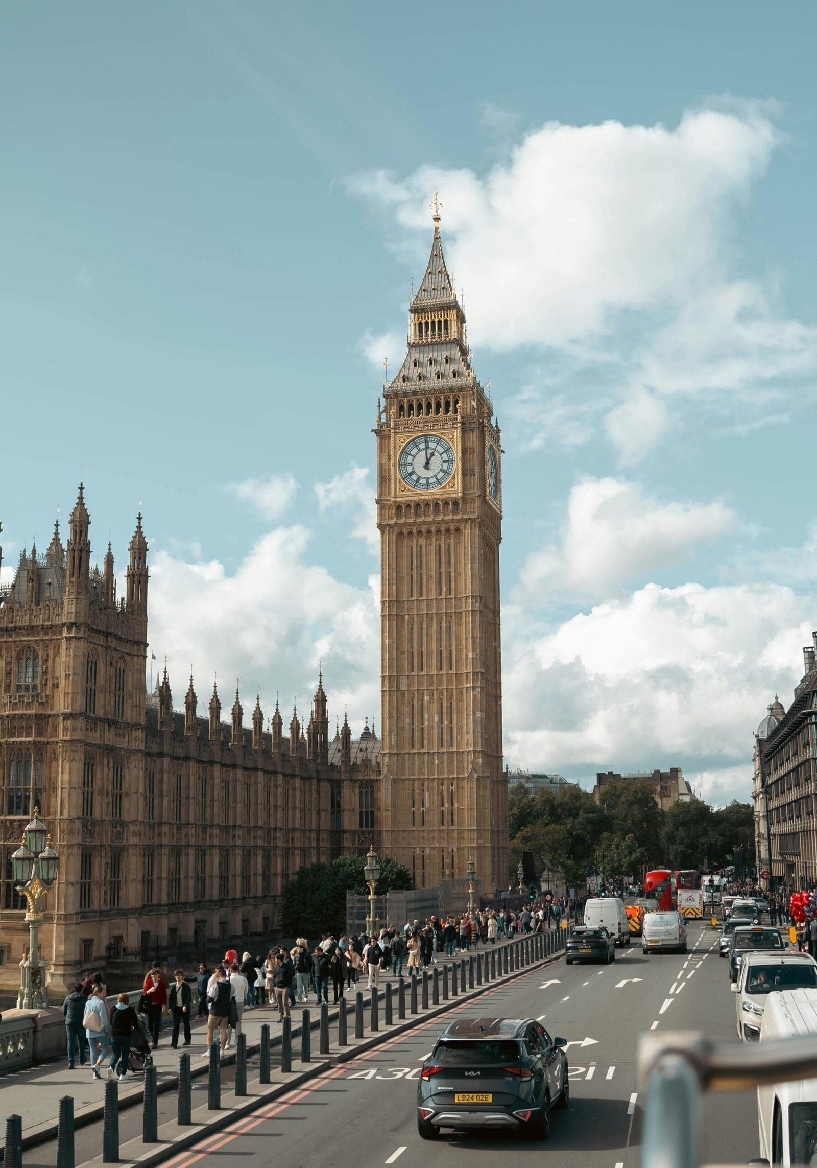 Monumento icónico de Londres, que es parte de su cultura. Se puede ver una torre de gran altura que tiene un reloj en la parte superior con cuatro esferas, una en cada lado de la torre. La torre parece ser de piedra de color claro con increíbles detalles arquitectónicos. Hay mucha gente caminando por la banqueta y un poco de tráfico en la avenida. Y se ve un clima caluroso por los rayos de sol que iluminan la ciudad.