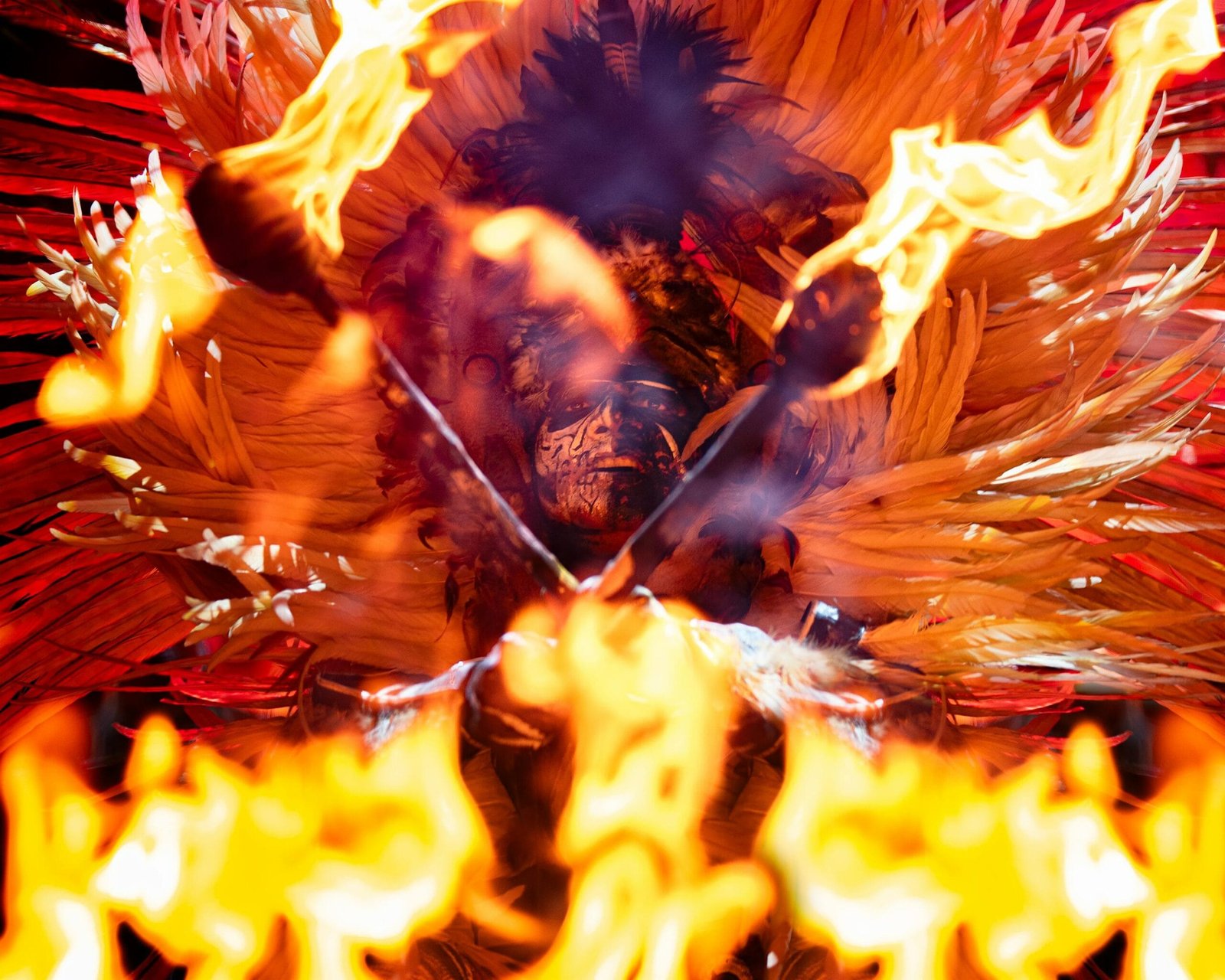 Una persona vestida con atuendo típico lleno de plumas de colores intensos, especialmente rojos, naranjas y amarillos está rodeada de fuego y sostiene antorchas encendidas. El fuego y las plumas crean un efecto visual poderoso, casi místico, la persona tiene el rostro pintado como en una ceremonia o danza ancestral.