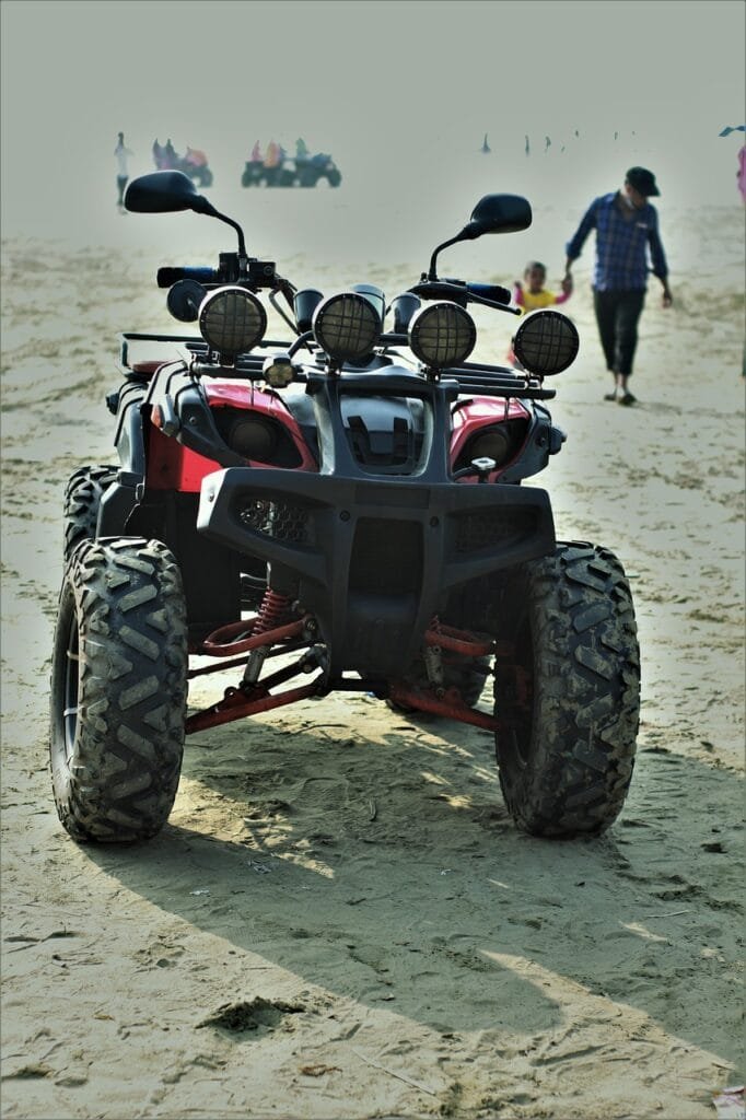 Un cuatrimoto todoterreno, estacionado sobre la arena en una playa, con personas caminando en el fondo. La imagen transmite aventura, libertad y actividades recreativas al aire libre.