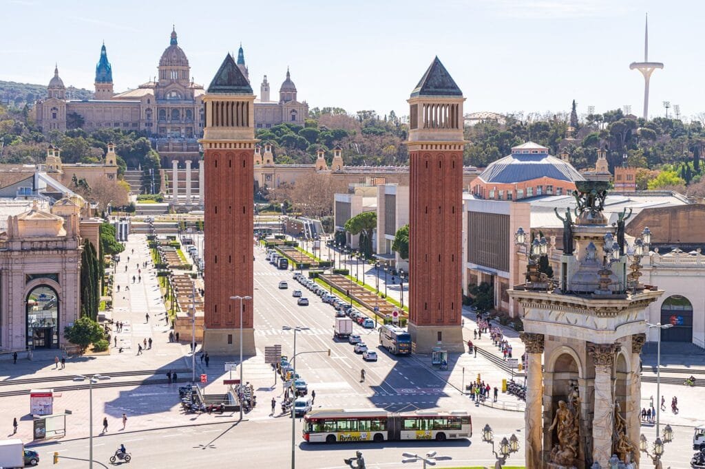 Se aprecian varios edificios con estilos arquitectónicos clásicos y modernos, también está presente la naturaleza como jardines y árboles. Se puede ver la fuente de la Plaza de España de lado derecho de la imagen, las Torres Venecianas al centro y el Palacio Nacional de Barcelona al fondo. Es de día, lo cual indica que el clima es caluroso. Hay poco flujo de gente y de automóviles, así como transporte urbano. La avenida que se extiende entre las torres Venecianas y el Palacio Nacional es utilizada frecuentemente para ferias, eventos y exposiciones. También se pueden encontrar tiendas y restaurantes a lo largo de la avenida formando parte de la cultura de Europa.
